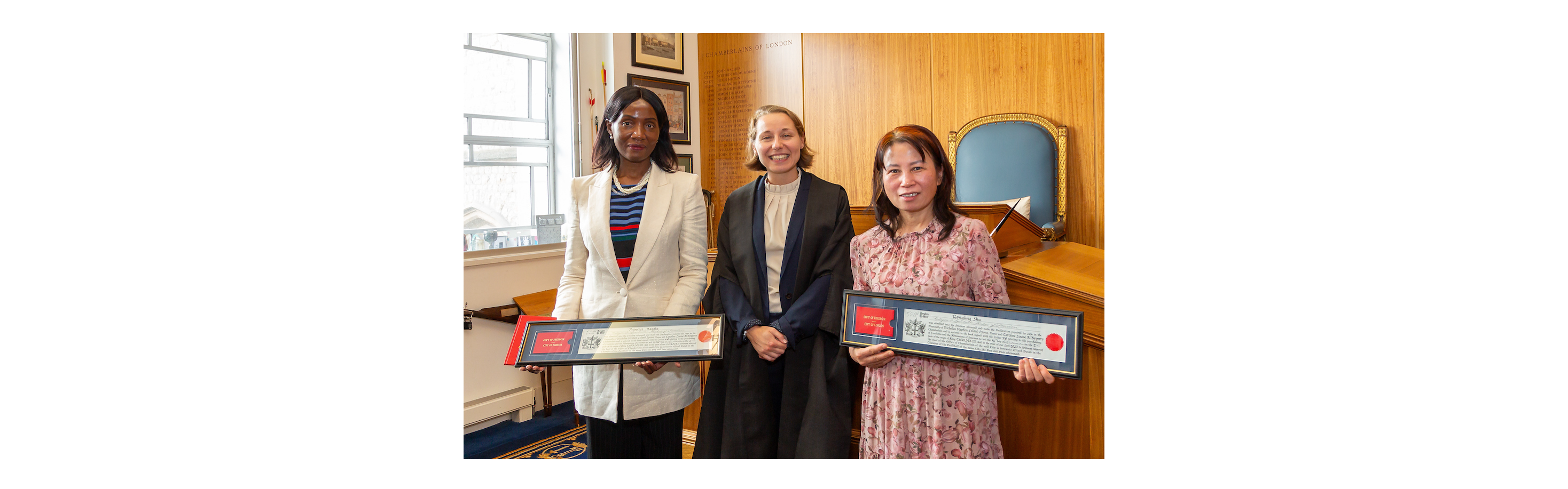 Image shows two Ophthalmic Nurses receiving their Freedom of the City of London as Spectacle Makers. They are holding their City Freedom certificates with an officer of the Chamberlain's Court.